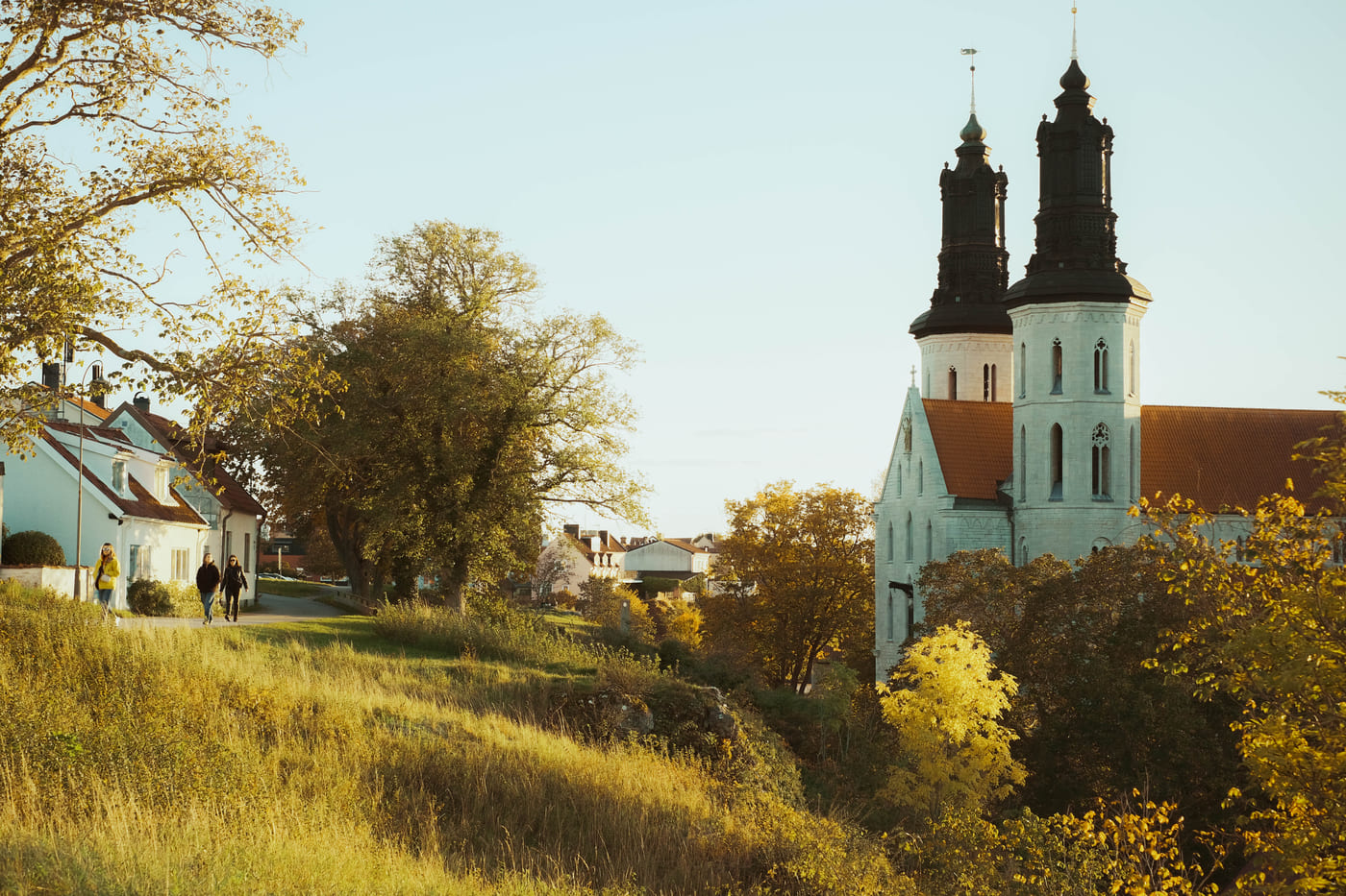 Visby cathedral, its two black spires rising above autumn trees, with people walking along a path below