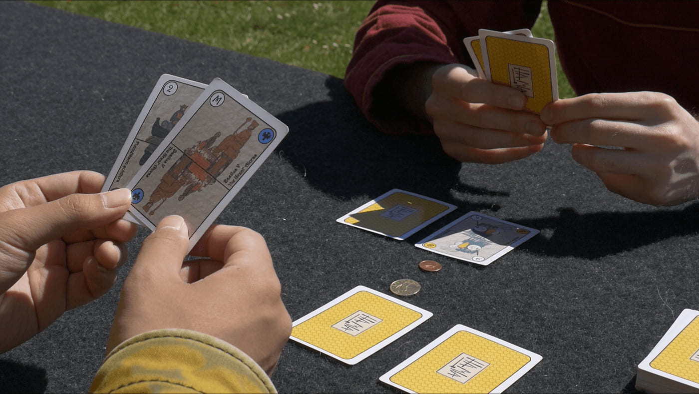 Two players holding illustrated Hamla cards outdoors, with yellow-backed cards laid out on a grey mat between them