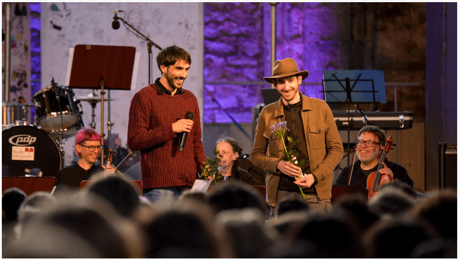 Two game designers on stage holding flowers at the Gotland Game Conference 2025 award ceremony, with musicians and an audience around them