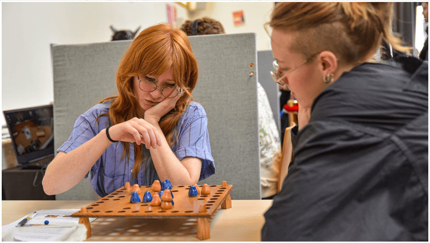 Two players facing each other across the Essom board, a wooden grid of holes holding blue and orange cone-shaped pieces